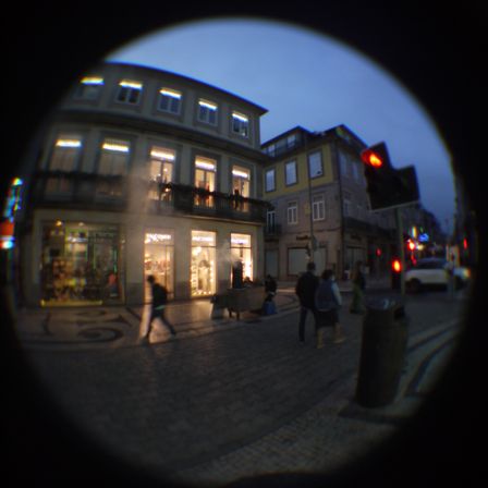 Foto de uma rua ao entardecer. O céu esta ficando escuro, tem poucas pessoas na rua e um prédio de três andares, a luz do térreo estoura na foto saindo de três portas