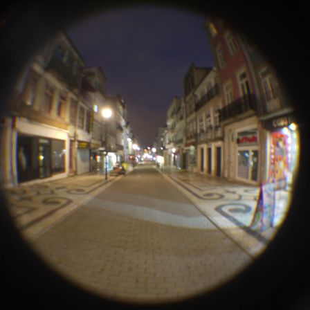 Foto de uma rua reta, a luz esta mais visivel, é possível reparar melhor na loja aberta ao lado direito, os prédios do lado esquerdo, mas a rua esta vazia.