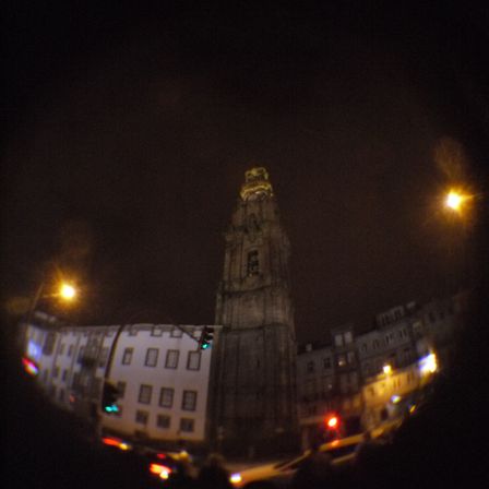 Foto no escuro de uma torre alta de uma igreja, em volta alguns prédios chegam apenas na metade do tamanho dela. O céu esta escuro ao fundo.