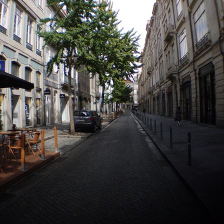 Foto de uma rua, vários prédios de 5 andares ao lado, duas árvores do lado esquerdo e na direita uma calçada com apenas uma pessoa. Na esquerda mesas de madeira em um deck ao lado da rua e mais a frente um carro estacionado.