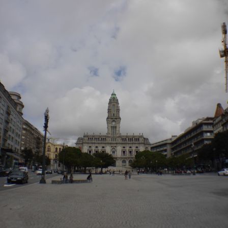 Foto de uma praça, quase sem árvores com uma rua a esquerda. Ao fundo um prédio antigo de 3 andares com uma torre no meio.