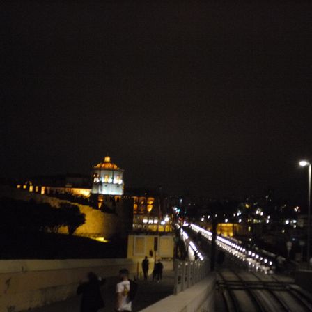 Foto de uma paisagem, esta a noite e as luzes destacam uma ponte. Ao fundo uma construção circular com um telhado pontudo e iluminado.