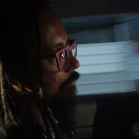 Foto em close e de perfil de uma pessoa preta (eu). Usando um óculos rosa e ao fundo um cenário embaçado e em movimento