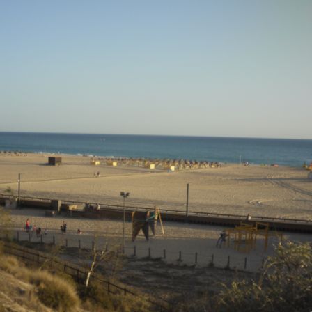 Foto de uma praia. É possível ver uma rua abaixo, com pessoas caminhando, a areia com algumas barracas eo mar ao fundo.