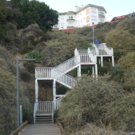 Foto de uma escada, ela sobe indo da direita pra esquerda por 2 andares e depois some entre as formações rochosas. Em cima da pra ver parcialmente um casarão.