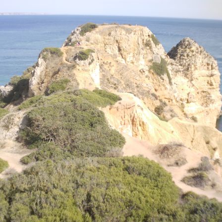 Foto de uma formação rochosa em formato de "pico", há vários caminhos até o topo. Ela ocupa boa parte do centro da foto e ao fundo da pra ver o mar e um pedaço do céu.