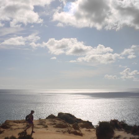 Foto do céu cheio de nuvens e o mar. Abaixo é possível ver um pedaço de terra e duas pessoas caminhando sozinhas.