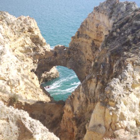 Foto de uma formação rochosa. A foto foi tirada de cima, ao fundo é possível ver o mar e há um arco no meio das rochas que da pra ver o mar e algumas ondas.