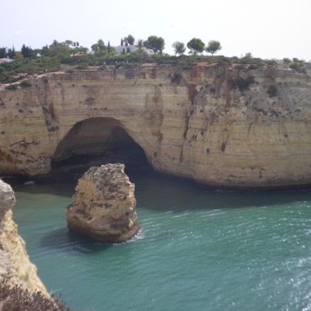 Foto do mar, ao fundo um penhasco com uma caberna no meio. Logo na frente da caverna tem uma pequena formação rochosa que quase cobre a entrada.