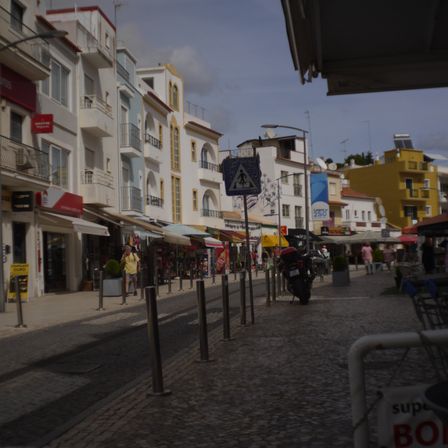 Foto de uma rua de asfalto e calçada portuguesa. No lado esquerdo várias casas de 3 andares, pintadas de branco com detalhes amarelos.